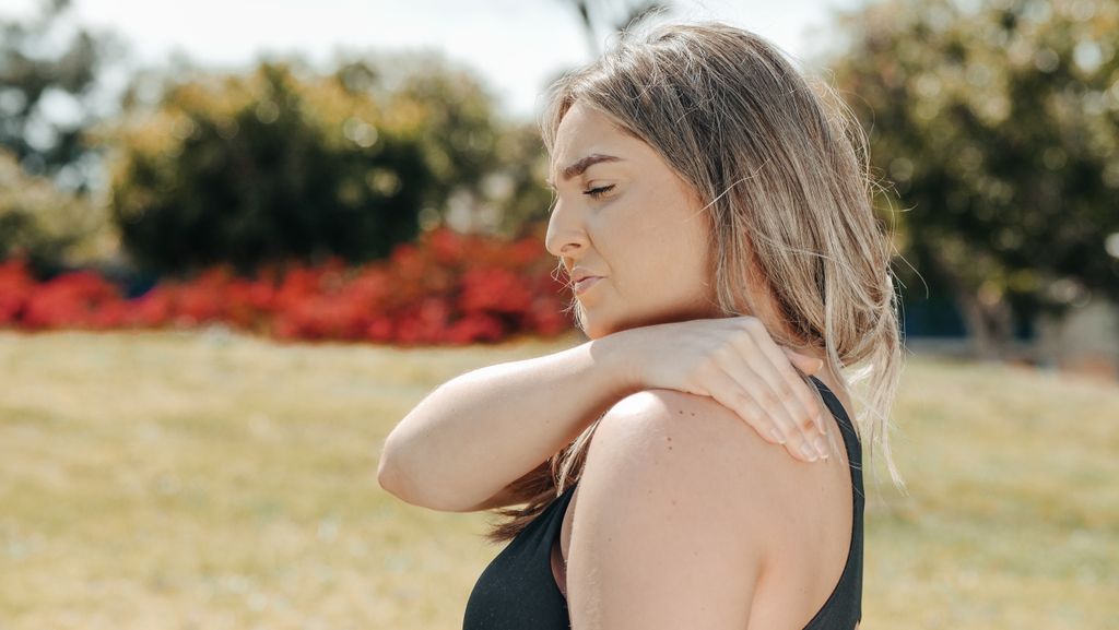 Woman in black tank top touching shoulder outdoors. Concept of shoulder pain and relaxation.