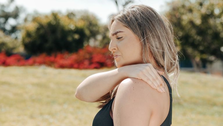 Woman in black tank top touching shoulder outdoors. Concept of shoulder pain and relaxation.